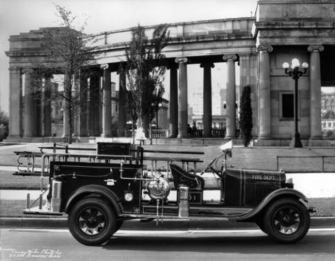 The Idaho Springs, Colorado, fire truck is in front of the Denver Civic Center. The truck is equipped with an axe, two fire extinguishers, ladders, fire hooks, a small, pressurized water container, a fire bell, a large head lamp mounted where the windshield would be, and a flashlight. There is a two person front seat, and running boards surround the body. "Idaho Springs Fire Dept.," is painted on the hood. Behind the truck, ionic columns accent Civic Center structures. (6-1-1929)